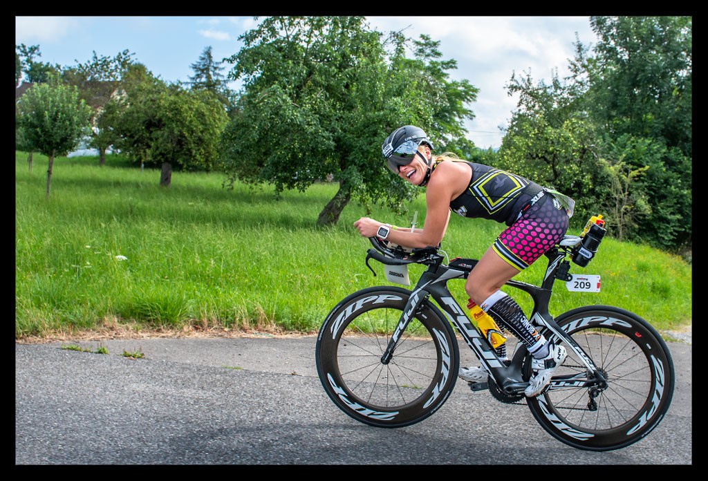 triathletin zeitfahrrad lächelt radstrecke ironman triathlon langdistanz farbiger tri-suit aero helm verspiegelt landschaft baum rasen sommer schweiz freude