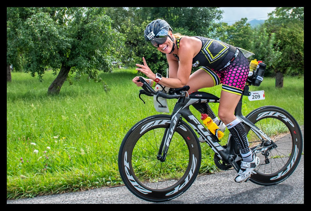 triathletin grüßt auf zeitfahrrad lächelt radstrecke ironman triathlon langdistanz farbiger tri-suit aero helm verspiegelt landschaft baum rasen sommer schweiz