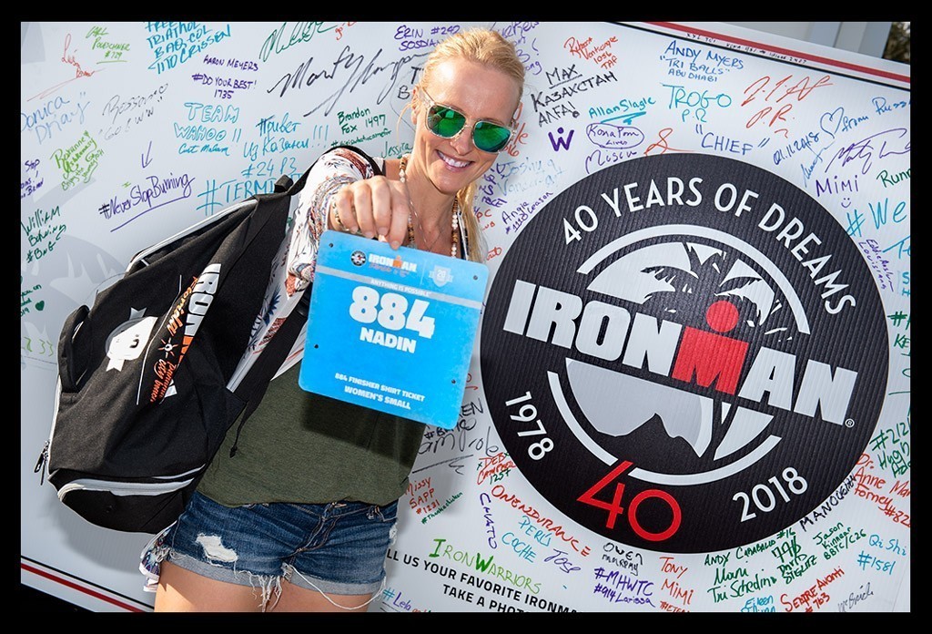Ironman Triathletin mit Hotpants Sonnenbille Rucksack zeigt Startnummer mit Logo Wand 40 Years of Dreams Ironman Florida 1978 2018 und Autogramme. Sie lächelt zufrieden