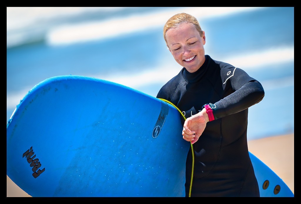 Frau mit Surfbrett am Meer blickt auf den Garmin Forerunner 645 und lächelt dabei.