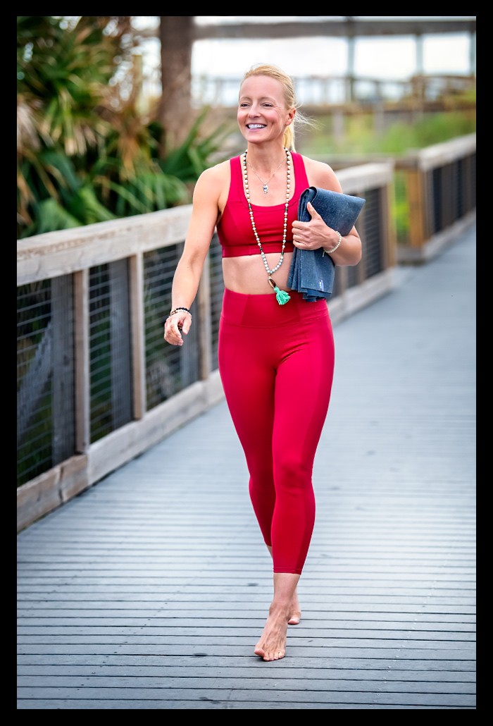Eine Blonde Frau in roten bauchfreiem Yoga-outfit läuft barfuß auf einem Steg und lächelt. Um den Hals eine lange Kette. In der Hand eine gefaltete Yogamatte.