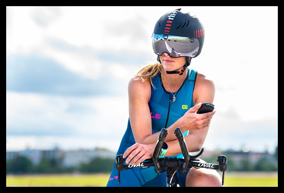 Triathletin mit einem Zeitfahrrad und Aerohelm. Sie hält das Garmin EDGE 1030 Plus GPS-Fahrradcomputer in der Hand. Sie trägt einen Tri-Suit von ALE in Hellblau mit der Garmin Forerunner 745 Multisportuhr am Handgelenk.