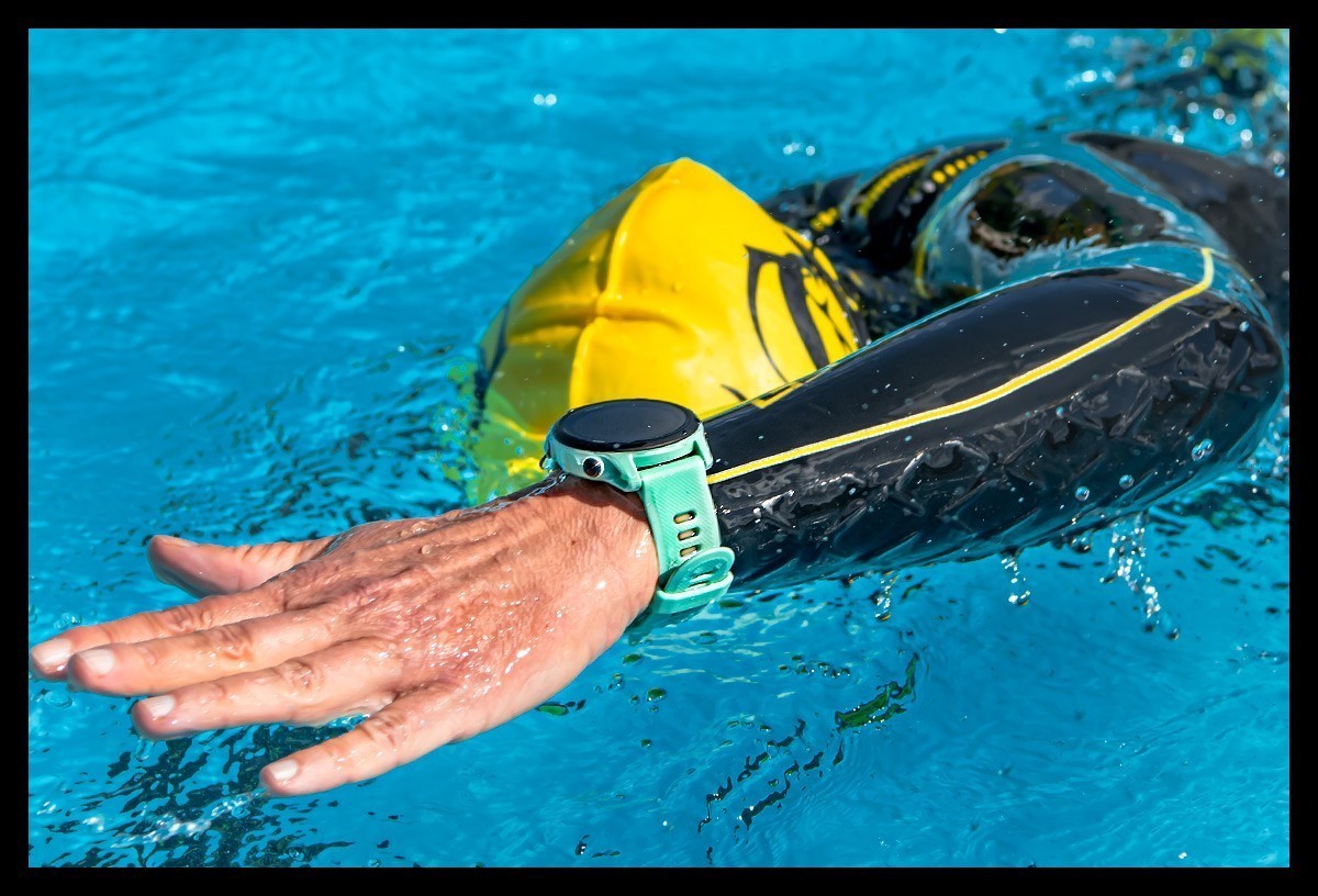 Triathletin schwimmt im blauem Wasser. Sie trägt einen Neoprenanzug von Sailfish, gelbe Badekappe, Schwimmbrille und Garmin Forerunner 745 Multisportuhr. Detaillierte Nahaufnahme von Hand und Uhr. Wasserspritzer. Produkttest. Review.