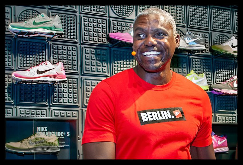 Carl Lewis Portrait Olympiasieger Nike Berlin besuch im Nike-Store mit rotem T-Shirt und Laufschuhen an der Wand lacht und zeigt Zähne glücklich