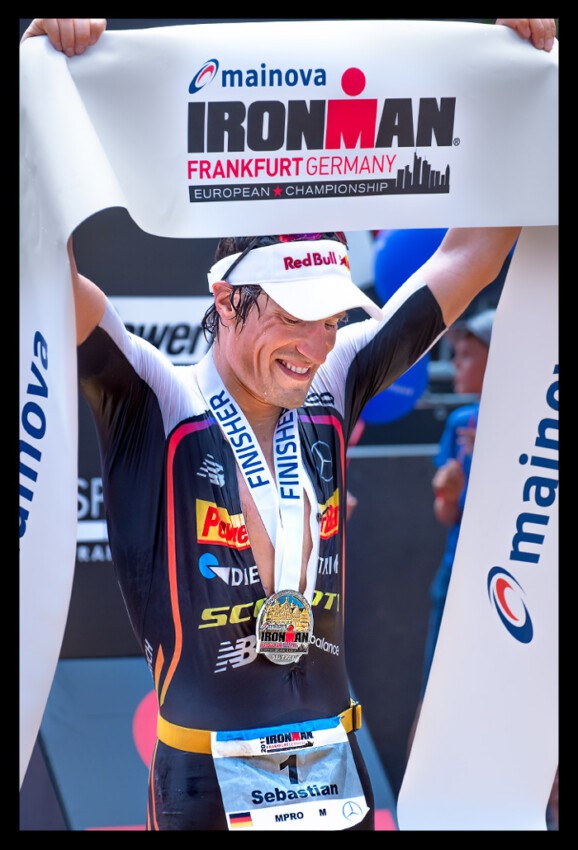 Sebastian Kienle Sieger IRONMAN Frankfurt Germany im Ziel mit Siegerschleife überm Kopf, Medailie um Hals und lacht glücklich