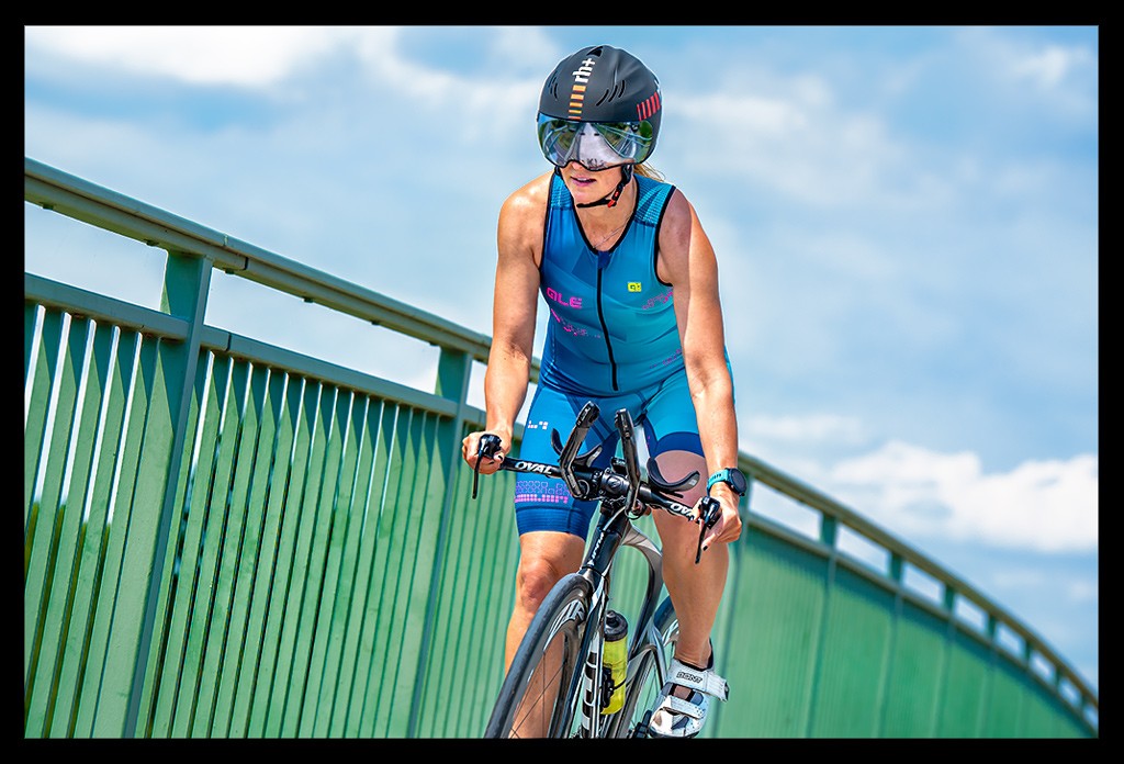 Triathletin im Tri-Suit mit Helm, Zeitfahrrad und BiSaddle Fahrradsattel für Triathleten und Radsportler auf Brücke vor blauem Himmel. Sie fährt im Wiegetritt und kippt das Rad zur Seite.