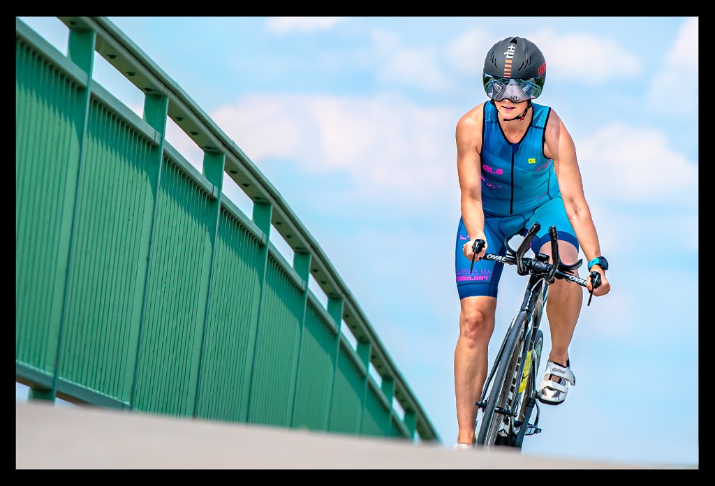 Triathletin im Tri-Suit mit Helm, Zeitfahrrad und BiSaddle Fahrradsattel für Triathleten und Radsportler auf Brücke vor blauem Himmel. Sie fährt im Wiegetritt und kippt das Rad zur Seite.