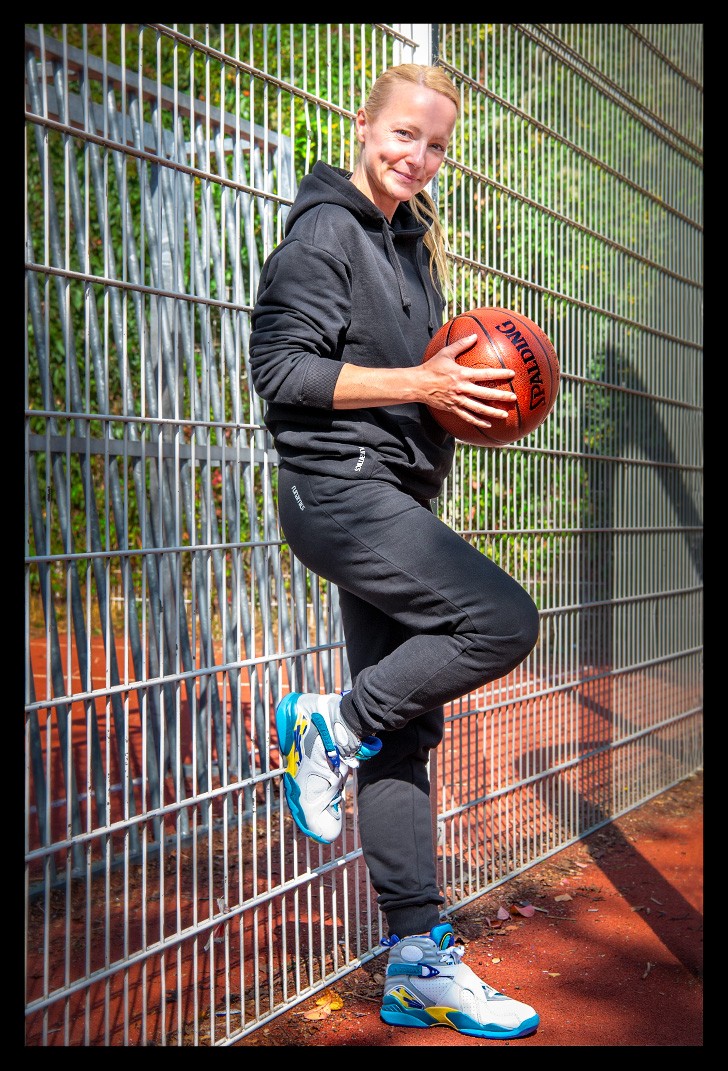 Nadin von eiswuerfelimschuh.de testet die neue Garmin beim Basketball spielen und hält den Ball mit beiden Händen. Sie lächelt und steht an einem Metallzaun auf einem Basketball Court.