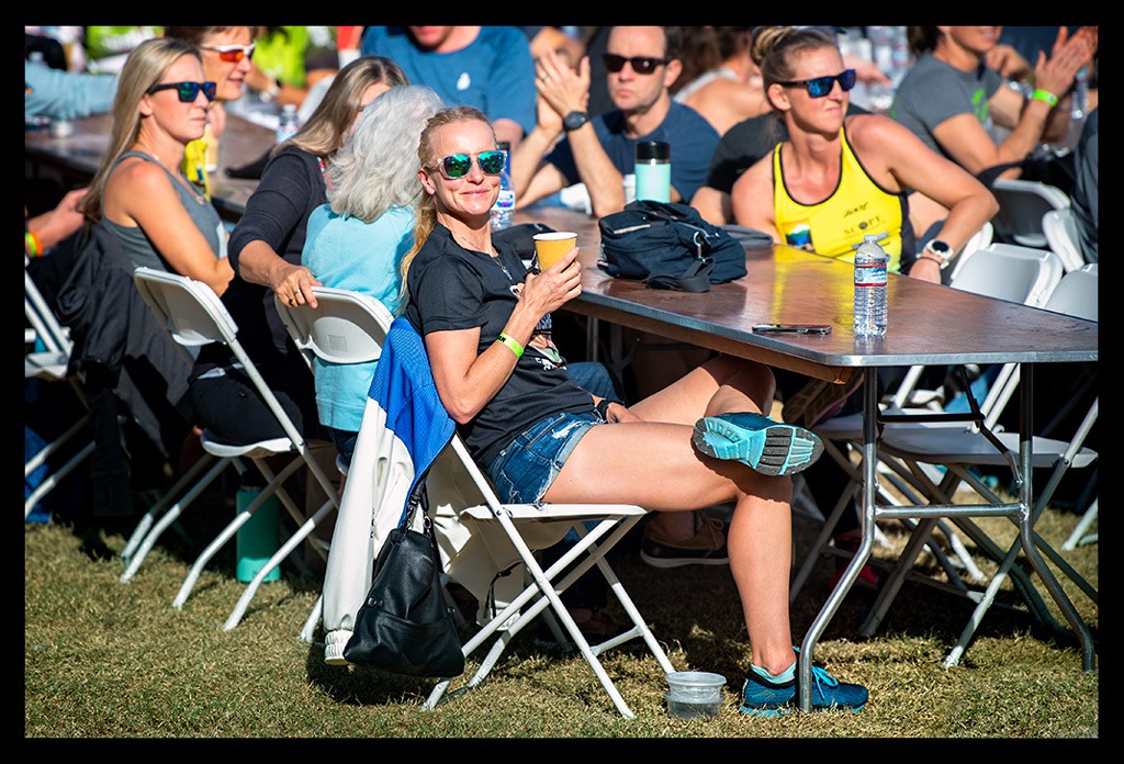 Nadin Eule Bloggerin und Triathletin von eiswuerfelimschuh.de bei der siegerehrung award ceremony Ironman Arizona Tempe USA. Sitzt am Tisch mit sonnenbrille und wasserflasche sowie kaffebecher. Andere Zuschauer im Hintergrund.Sommerliche stimmung