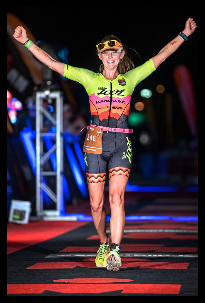 women triathlete finishline Ironman-Arizona-Tempe  USA flashlights red carpet "M" Logo Ironman celebrating holding arms up and smiles