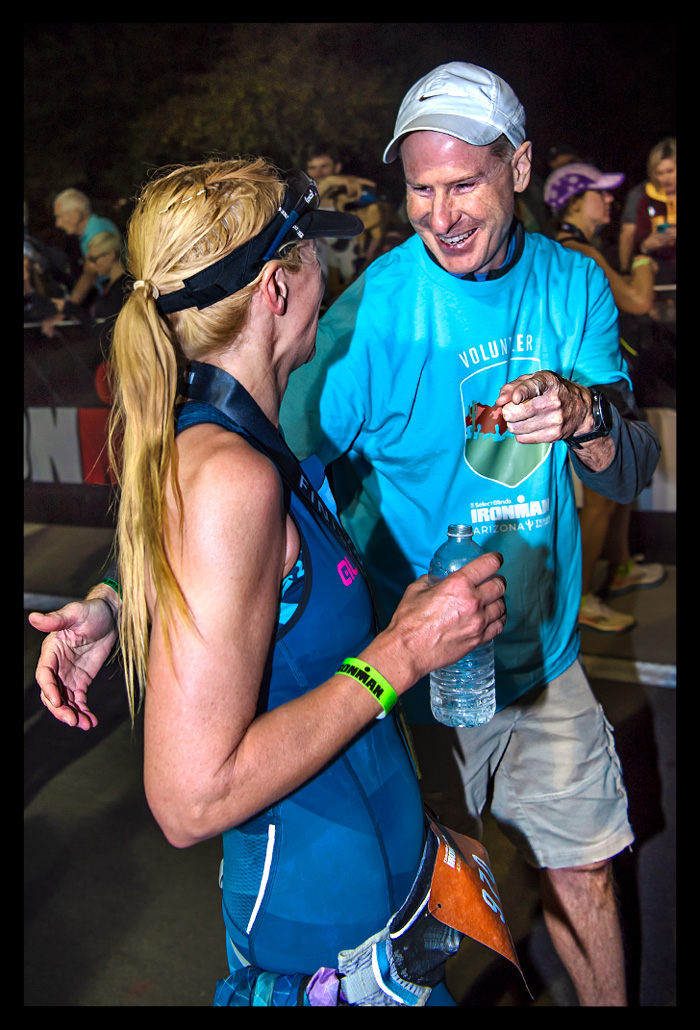 Nadin im Ziel Ironman Arizona Tempe Phoenix USA nach erfolgreicher Langdistanz mit lächeln im Gesicht und Helfer an der Seite mit Medaille um den Hals. Helfer zeigt auf Fotografen Oliver und lacht freudig