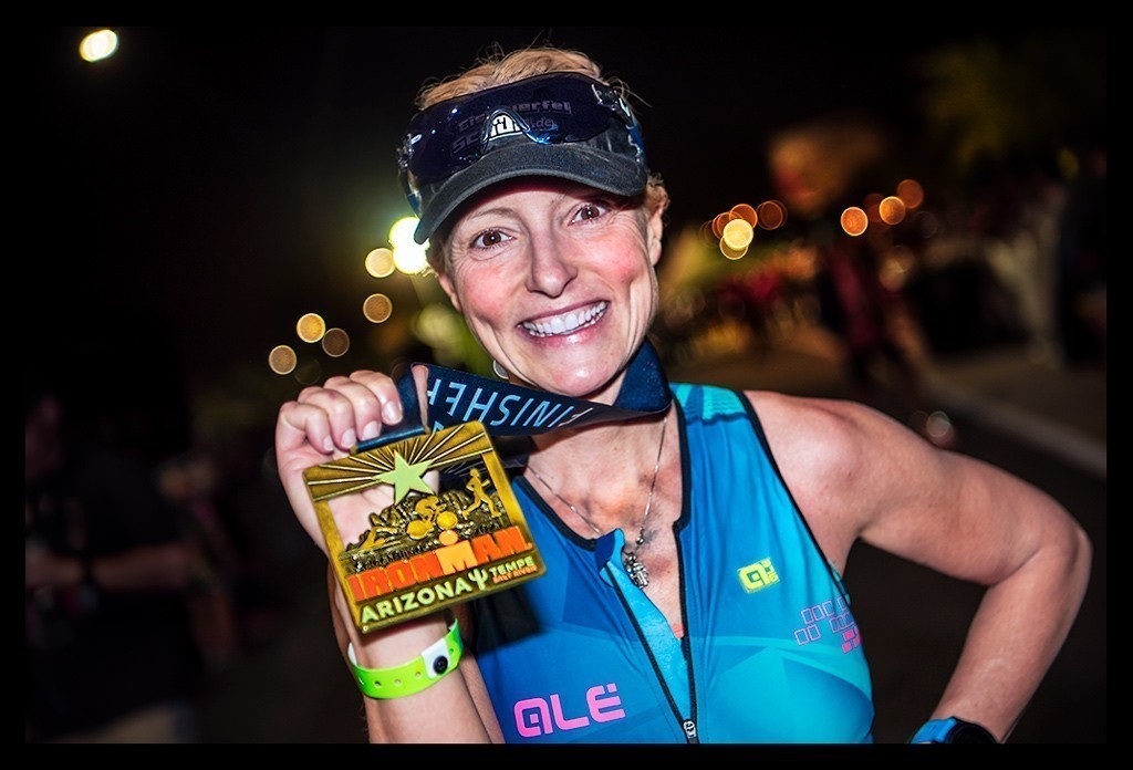 Ironman Arizona Tempe USA, Nadin Bloggerin und Triathletin von eiswuerfelimschuh.de zeigt lachend ihre Finisher Medaille. Blauer Tri-Suit Garmin Forerunner in abendstimmung mit laternen lichtern im hintergrund. Medaille ist riesig