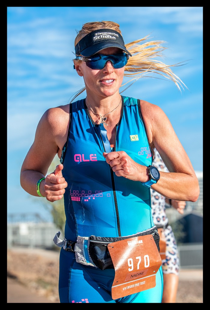 Ironman Arizona Nadin Bloggerin und Triathletin auf der Laufstrecke bei Sonnenschein und blauem Himmel mit blauen Tri-Suit von "ALE" und Garmin Forerunner. Startnummer 970. Sie lächelt und blonder Pferdeschwanz bewegt sich im Wind