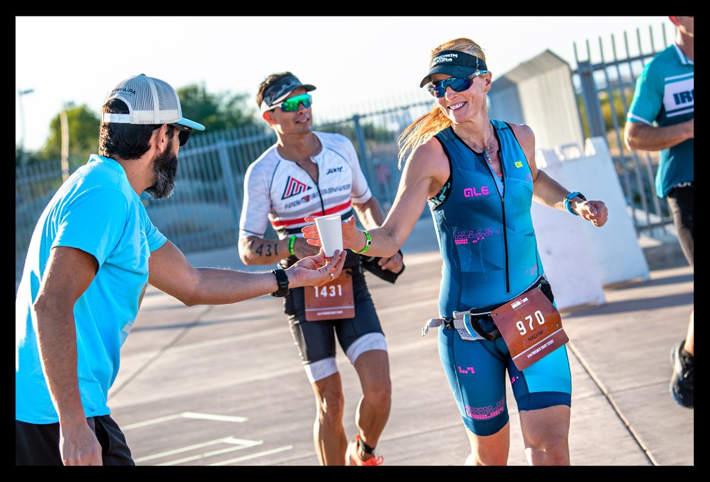 Nadin Bloggerin und Triathletin von eiswuerfelimschuh.de auf der Laufstrecke Ironman Arizona Tempe an Verpflegungsstation bekommt von Helfer Wasserbecher gereicht.sie läcjhelt im blauen Tri-Suit mit Garmin Forerunner und Startnumme 970. Athleten im Hintergrund