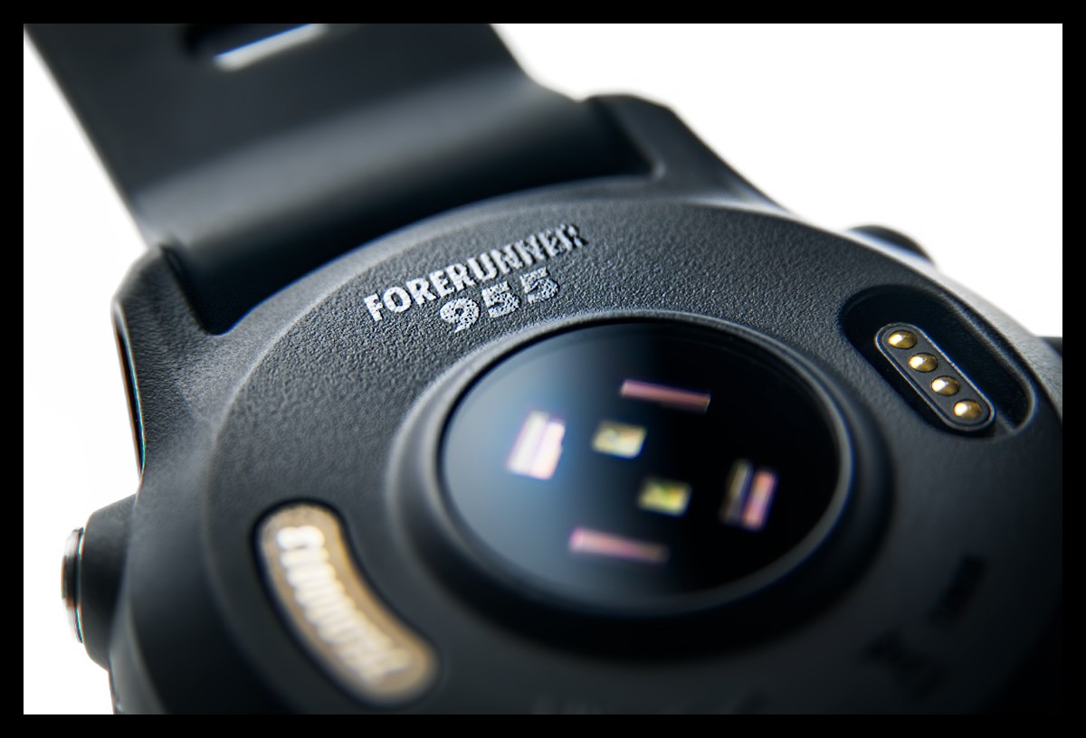 Garmin Forerunner 955 Multisportuhr GPS detaillierte Nahaufnahme Unterseite vom PulseOx und HF Sensor sowie Metall Tasten Button und Anschluss Ladekabel. Schriftzug Forerunner 955 auf Gehäuse zu sehen mit Armband in schwarz. Glatte Oberfläche angeleuchtet