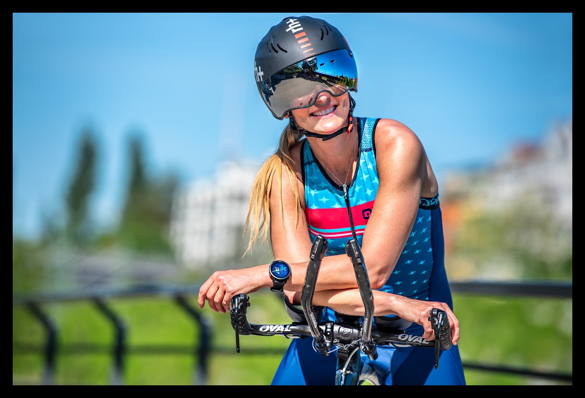 Triathletin mit dem Garmin Forerunner 955 Multisportuhr im Test und Fuji Zeitfahrrad TT Bike posiert vor Skyline und Geländer. Sie lächelt und trägt einen Helm von RHplus und Tri-Suit von ALE in sommerlicher Stimmung. Arme abgelegt auf den Lencker und lächelt im Kamera