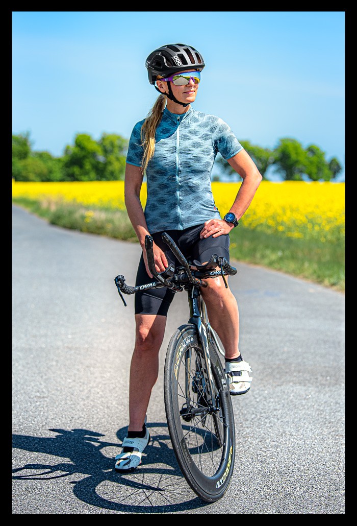 Frau Radsport posiert zeitfahrrad triathlon helm MIPS system poc sports octal schwarz garmin multisportuhr 955 strasse blumenfelder bäume landschaft sommerlich entspannt