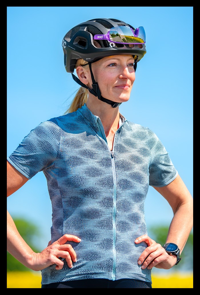 Frau Radsport Triathlon posiert hände in hüfte lächelt sommerlich blaues shirt landschaft bunt wiese blumenfelder bäume helm MIPS system poc sports octal schwarz garmin multisportuhr 955