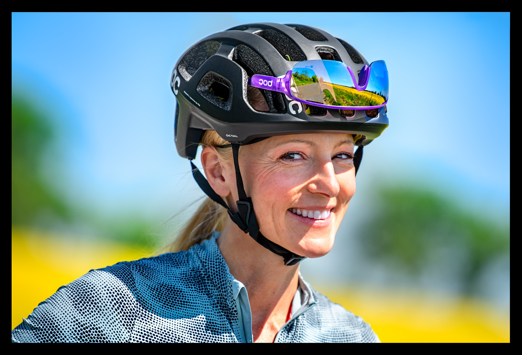 Radsportlerin Frau Portrait Fahrradhelm MIPS system Sicherheit lebensretter TEST POC sports lächelt blaues shirt landschaft bunt wiese blumenfelder bäume sommerlich