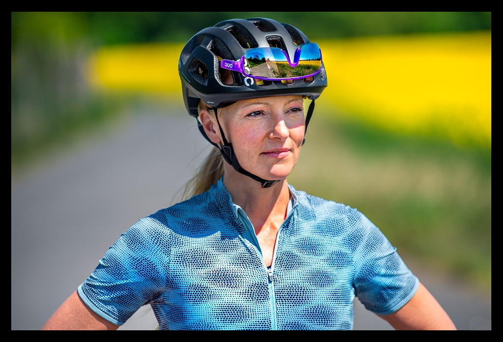 Frau Radsport Triathlon posiert hände in hüfte  sommerlich blaues shirt landschaft bunt wiese blumenfelder bäume helm MIPS system poc sports octal schwarz