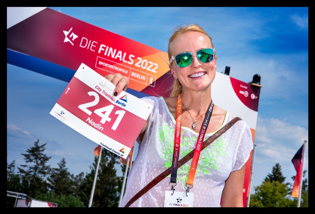 triathletin teilnehmerin city triathlon finals berlin zeigt startnummer vor zielbogen olympischer platz weiße bluse sommerlich himmel blau 