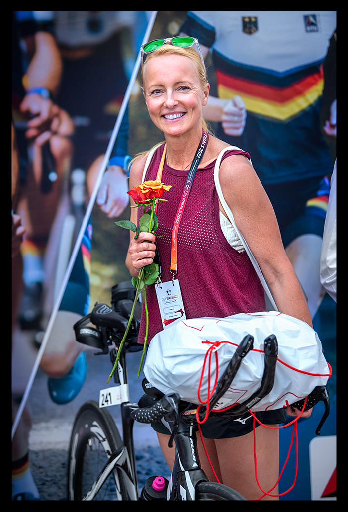triathletin nadin eule siegerehrung die finals berlin 2022 city triathlon sprintdistanz posiert mit zeitfahrrad und rose in der hand vor dtu plakat sie lächelt glücklich rotes oberteil kurze hose 