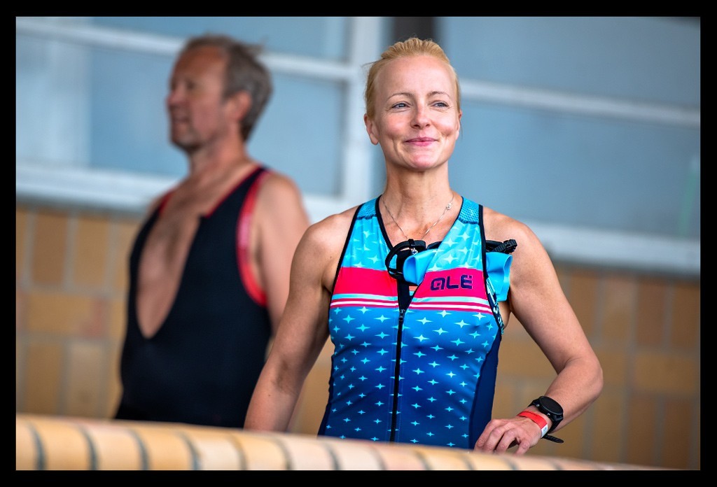 triathletin blonde haare blauer tri-suit ale cycling rennanzug lächelt vor schwimmstart steht strandbad wannsee blickt auf wasser berlin die finals 2022 city triathlon sprintdistanz