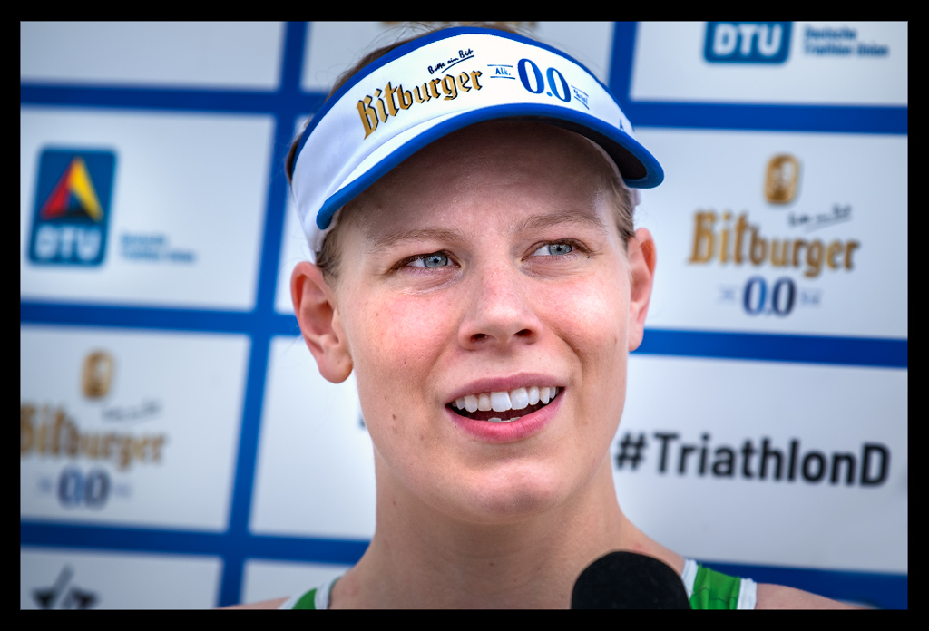 Laura Lindemann siegerin im tv interview lächelt die finals berlin deutsche triathlon meisterschaft sprintdistanz elite damen DTU vor werbebanner trägt 0.0% cap grüner rennanzug Triathlon Potsdam e.V. portrait nahaufnahme mit mikrofon