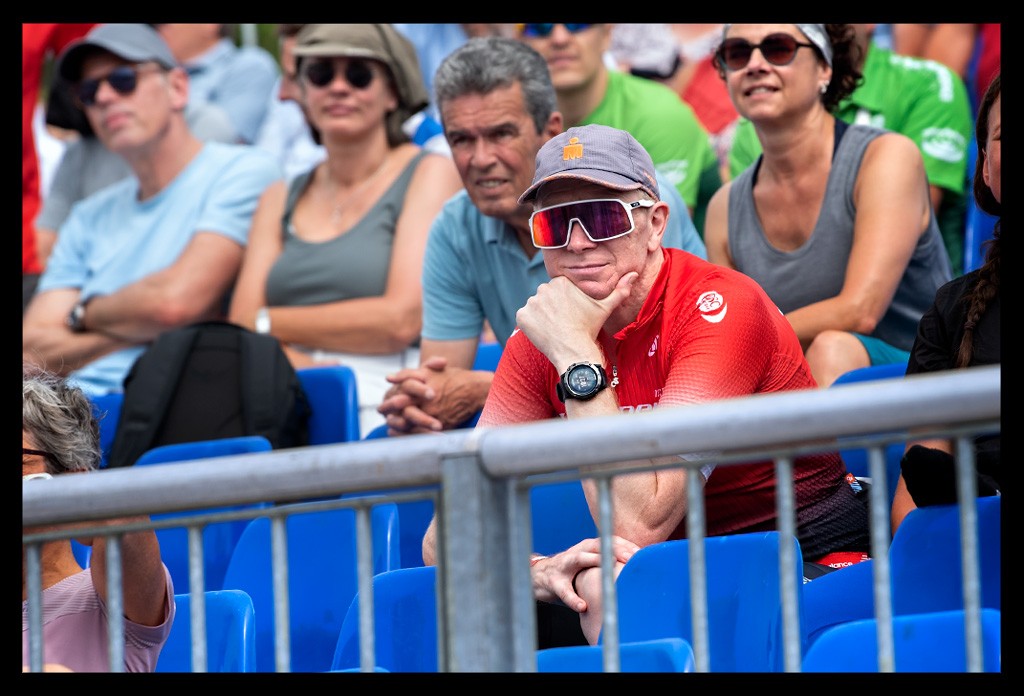 zuschauer auf tribüne bei die finals berlin deutsche DTU Triathlon meisterschaften sprintdistanz elite sommerlich warm blaue sitze mann trägt ironman cap mütze sonnenbrille blickt auf videowand