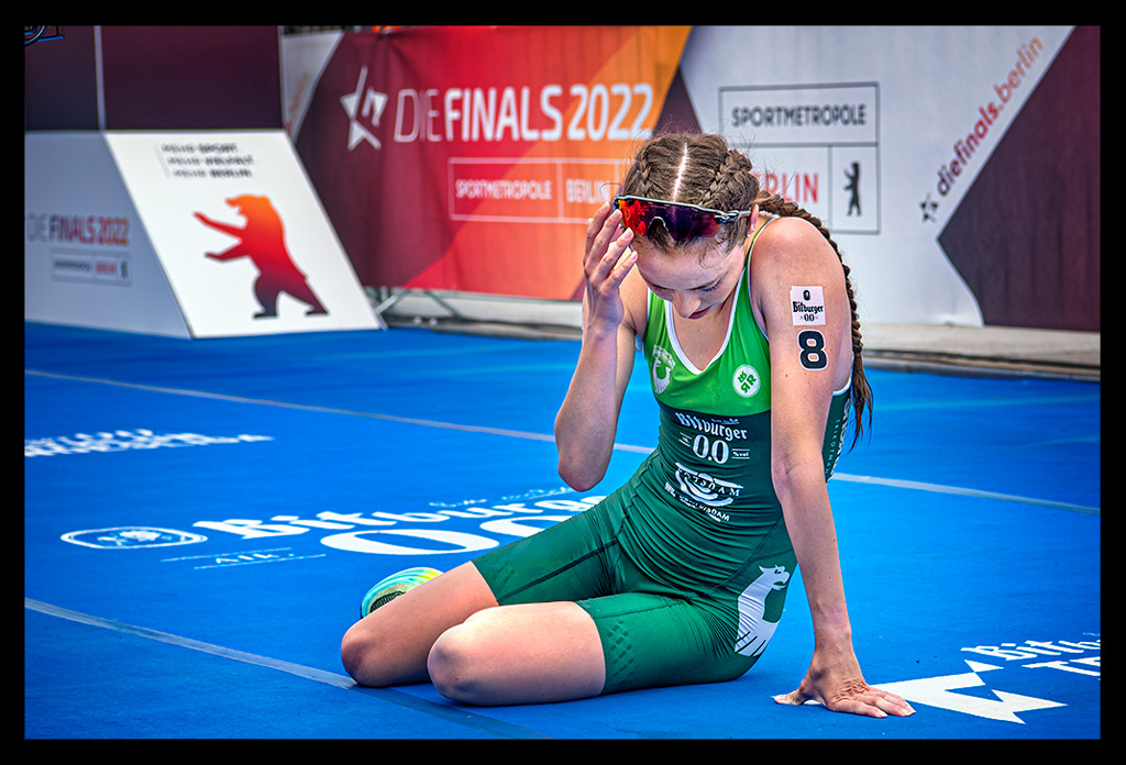 Selina Klamt erschöpft im ziel die finals berlin deutsche DTU triathlon meisterschaft sprintdistanz sitzt am boden grüner rennanzug Triathlon Potsdam e.V. blauer teppich werbebanner sie greift an stirn