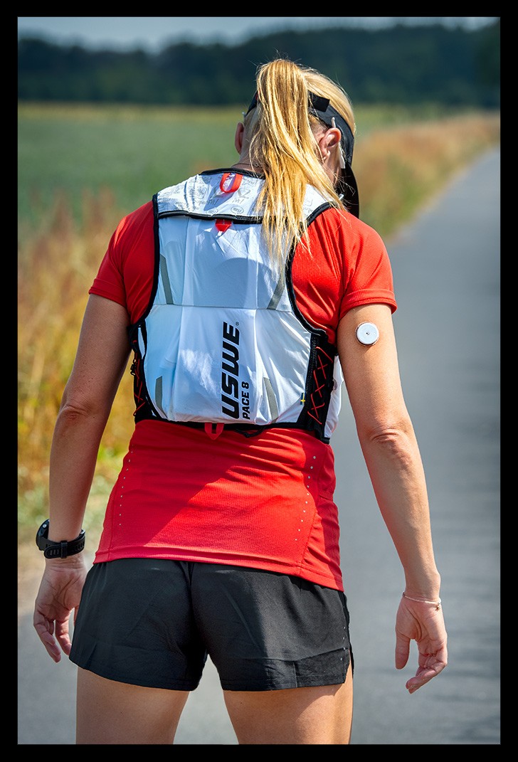 athletin posiert bei training mit Supersapiens Glucose-Biosensor abbott libre am arm blonde haare rotes shirt kurze schwarze hose Trailrunningrucksack USWE Pace Laufweste Getränke