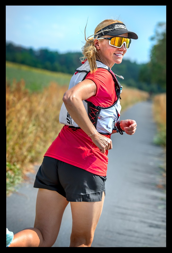 Läuferin trainiert auf feldweg blonde haare rotes shirt kurze schwarze hose mit Supersapiens Glucose-Biosensoren am arm überwacht Blutzuckerwerte und lächelt sommerlich warm