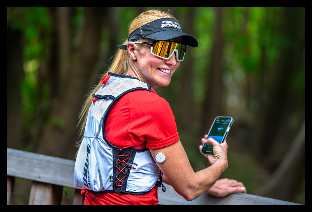 athletin überwacht Blutzuckerwerte mit Supersapiens Glucose Biosensor beim sport training blonde haare rotes shirt posiert auf brücke smartphone mit datenfelder app sie lächelt sommer wald