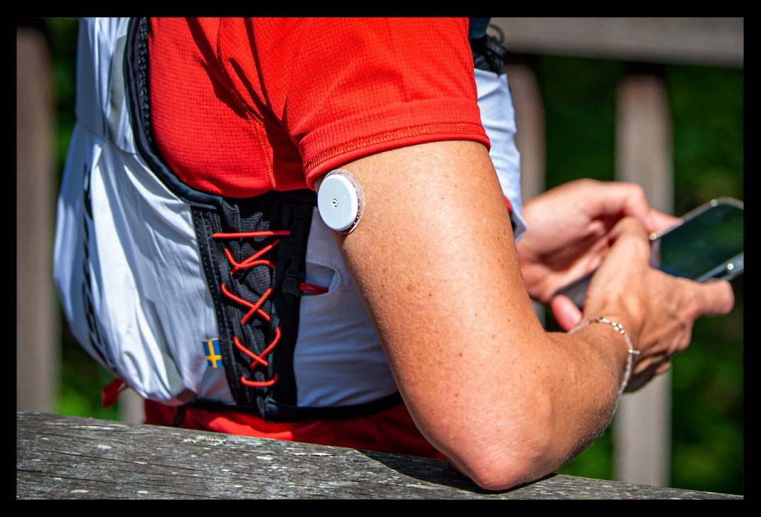 athletin überwacht Blutzuckerwerte mit Supersapiens Glucose Biosensor abbott libre smartphone mit datenfelder sensor mit nadel steckt im arm USWE Pace Laufweste