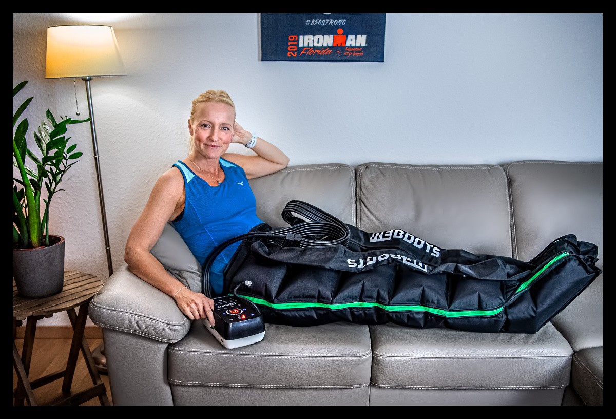 Blogger und Triathletin auf Couch mit Reboots Go Recovery Pants Boots zur Regeneration im Tank Top lächelnd, im Hintergrund ein Ironman Florida Poster an der Wand, eine Stehlampe leuchtend und eine Pflanze