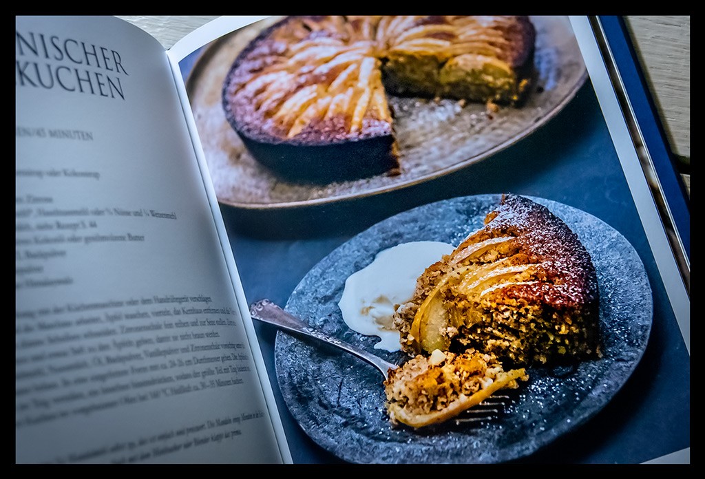 Buch "Nuss-Küche: Gesunde und reichhaltige Rezepte für Herzhaftes und Süßes" aufgeschlagen mit Foto von Apfelkuchen auf weißem Holzschreibtisch liegend