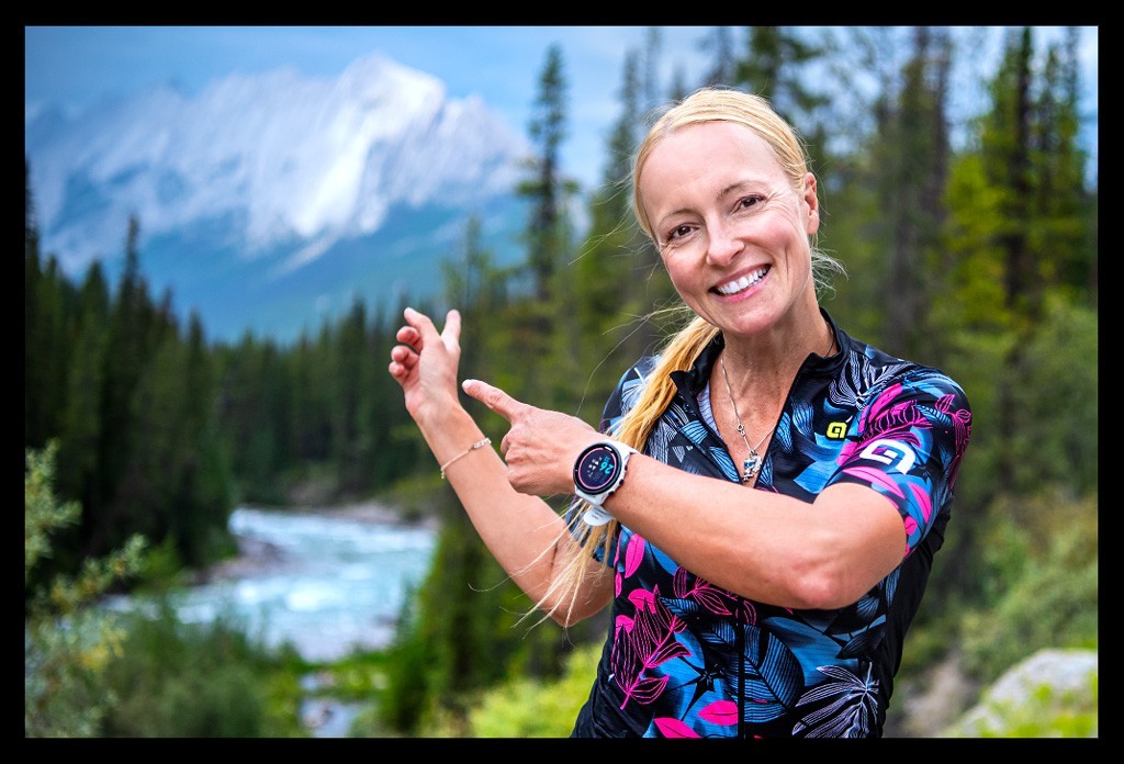 triathletin blonder zopf Radtrikot geöffnet lächelt posiert in den bergen von kanada mit landschaft im hintergrund mit Garmin forerunner 955 solar am handgelenk