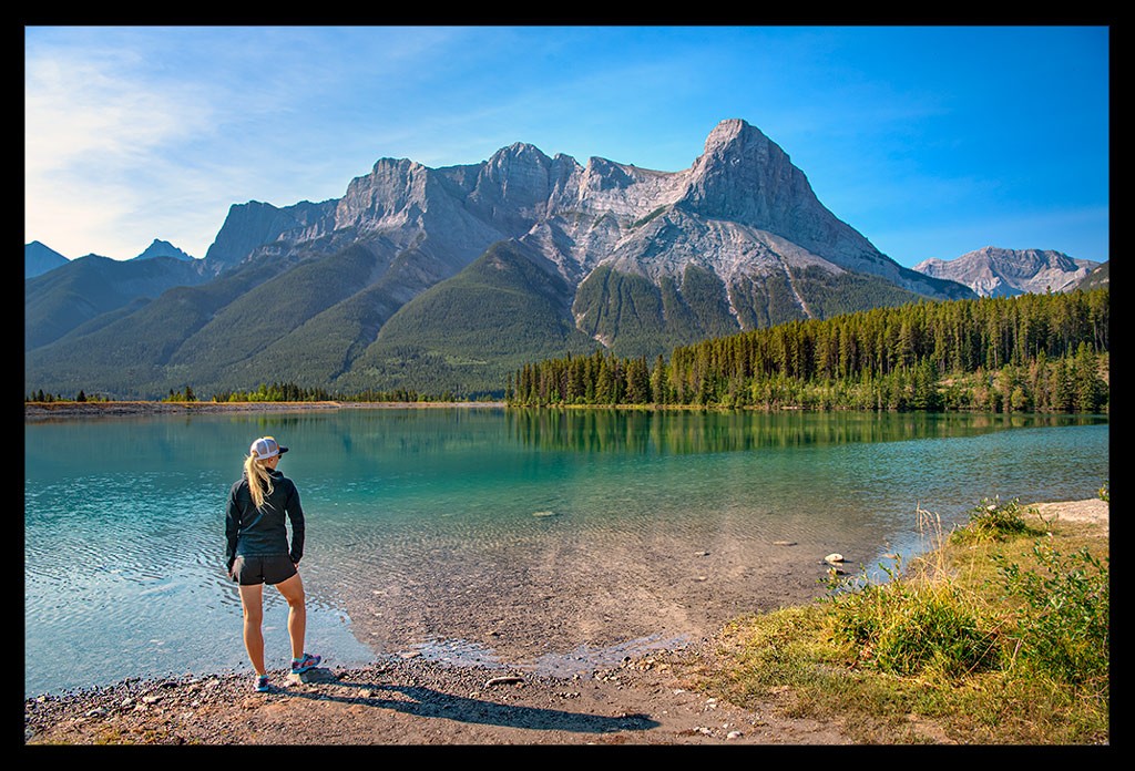 Kanada Road Trip durch die Rocky Mountains - hier Bloggerin Nadin an einem türkisfarbenem See stehend mit Blick auf diesen am Steinstrand, im Hintergrund hohe Berge und Nadelwälder