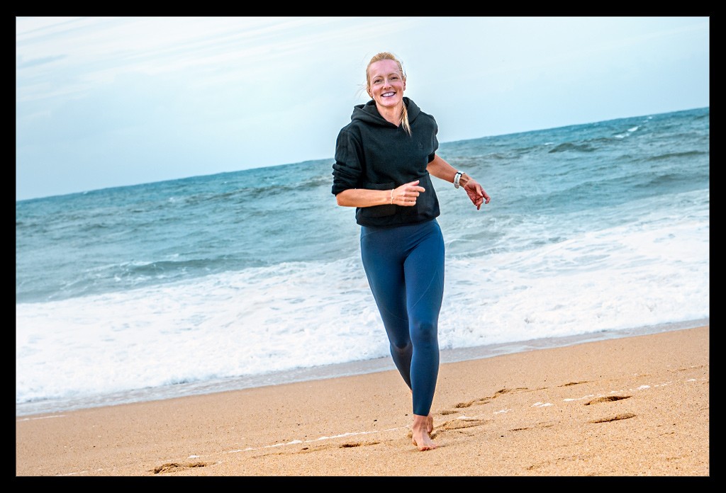 Blonde Frau läuft auf Strand am Atlantik entlang mit blauer Tights und schwarzem Hoodie für den Artikel über: Blue Mind und Blue Spaces - Glück am Wasser - erfahre, welchen positiven Einfluss die Nähe zum Wasser und Blue Spaces auf unser Wohlbefinden haben.