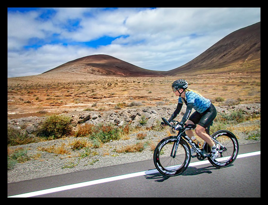 Frau mit Triathlonrad auf Lanzarote Kanaren mit Gebirge und Vulkangestein im Hintergrund. Blaues Rapha Outfit und Garmin Forerunner