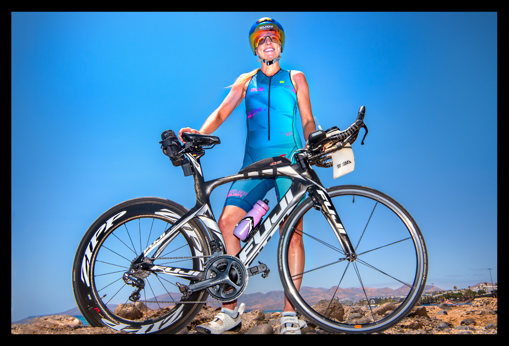 Triathletin lächelnd mit TriSuit und Aero-Helm hinter Zeitfahrrad auf Lavagestein und Sandstrand stehend - im Hintergrund blauer Ozean und Himmel