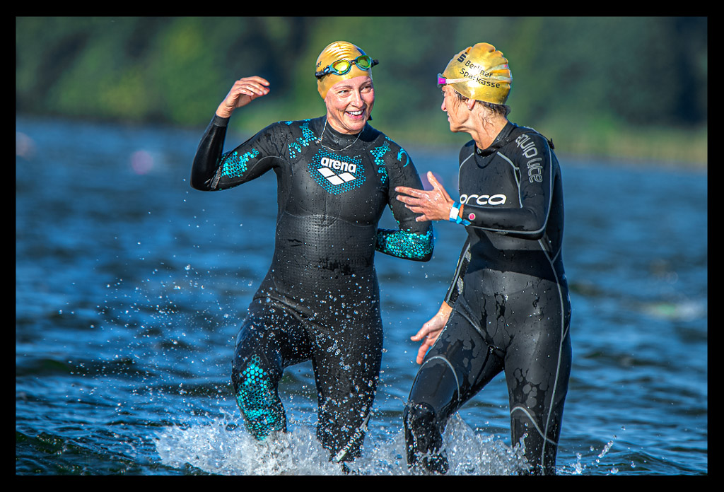 Triathletin Nadin von eiswuerfelimschuh.de lächelnd am Strandbad Wannsee mit einer anderen Athletin sprechend mit und Neoprenanzug an und goldener Jubiläums-Badekappe