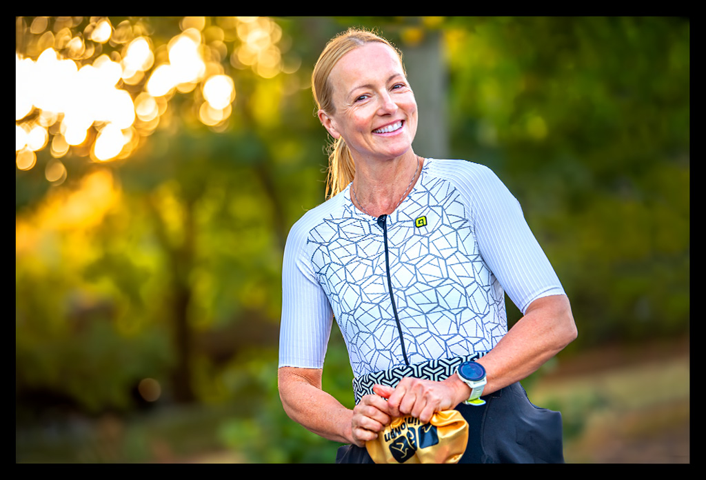 Triathletin Nadin von eiswuerfelimschuh.de lächelnd am Strandbad Wannsee in Morgensonne mit TriSuit und Neoprenanzug an und goldener Jubiläums-Badekappe in der Hand