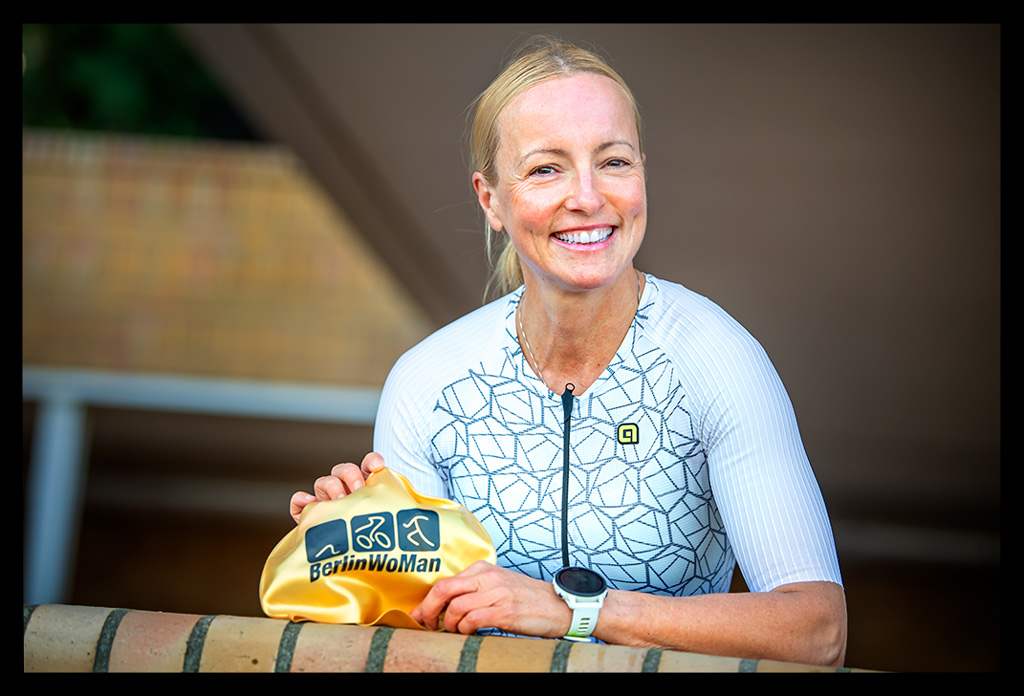 Triathletin Nadin von eiswuerfelimschuh.de lächelnd am Strandbad Wannsee in Morgens mit TriSuit und Neoprenanzug an und goldener Jubiläums-Badekappe in der Hand